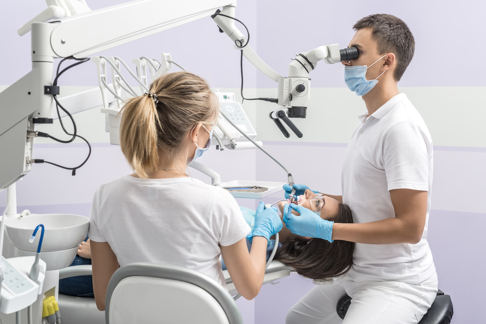 Woman in goggles on a dental chair and a dentist with assistant who sit next to her. Man looks on her teeth using a dental microscope and holds dental instruments. Blonde holds an air water syringe.